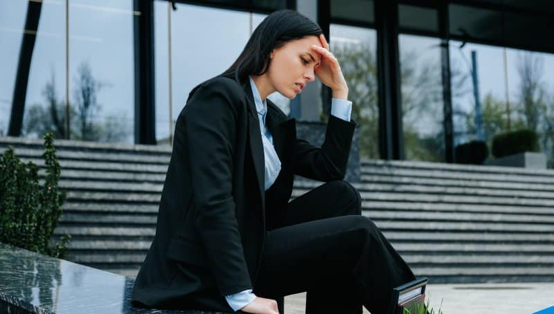 A woman is shown sitting outside office building looking upset and distressed.