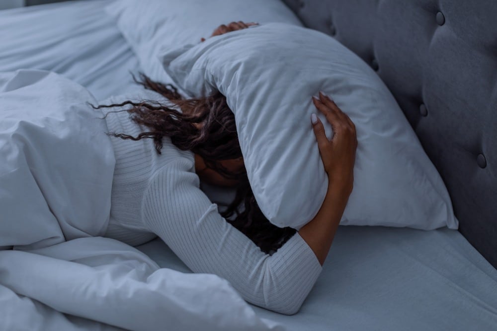 Stressed woman covering head under pillow lying in bed